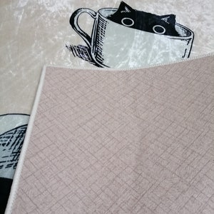 May include: A beige rug with a black and white illustration of a cat peeking out of a teacup. The rug has a textured, woven backing.