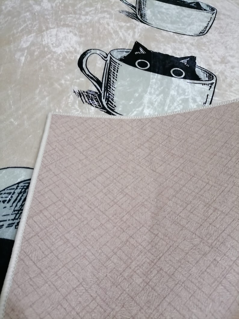 May include: A beige rug with a black and white illustration of a cat peeking out of a teacup. The rug has a textured, woven backing.