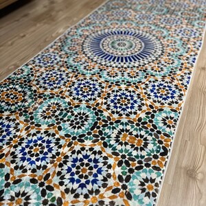 K&ouml;nnte beinhalten: Ein rechteckiger Teppich mit einem detaillierten geometrischen Muster in Blau-, T&uuml;rkis-, Braun- und Wei&szlig;t&ouml;nen. Das Design weist ein zentrales kreisf&ouml;rmiges Motiv und sich wiederholende sternartige Formen auf. Der Teppich liegt auf einem Holzboden.