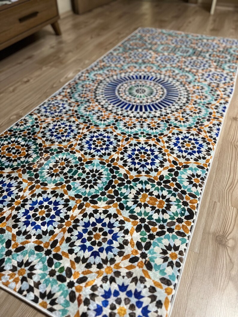 K&ouml;nnte beinhalten: Ein rechteckiger Teppich mit einem detaillierten geometrischen Muster in Blau-, T&uuml;rkis-, Braun- und Wei&szlig;t&ouml;nen. Das Design weist ein zentrales kreisf&ouml;rmiges Motiv und sich wiederholende sternartige Formen auf. Der Teppich liegt auf einem Holzboden.