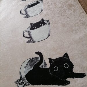 May include: A beige rug with a black cat illustration. The cat is depicted in various stages of entering a coffee cup. The cat is peeking out of the cup, then half-way in, and finally fully inside the cup.