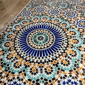 K&ouml;nnte beinhalten: Rechteckiger Teppich mit detailliertem geometrischem Muster. Das Design zeigt ein sich wiederholendes Mosaik aus blauen, t&uuml;rkisfarbenen, braunen und schwarzen Formen. Der Teppich liegt auf einem hellen Holzboden, das Muster ist in Nahaufnahme zu sehen.