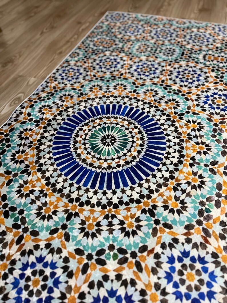 K&ouml;nnte beinhalten: Rechteckiger Teppich mit detailliertem geometrischem Muster. Das Design zeigt ein sich wiederholendes Mosaik aus blauen, t&uuml;rkisfarbenen, braunen und schwarzen Formen. Der Teppich liegt auf einem hellen Holzboden, das Muster ist in Nahaufnahme zu sehen.