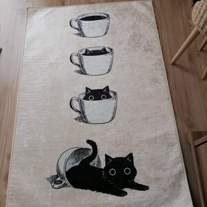 May include: A beige rug with a black cat peeking out of a coffee cup. The cat is lying on its belly with its paws outstretched. There are three coffee cups stacked on top of each other, with the cat peeking out of the middle cup.