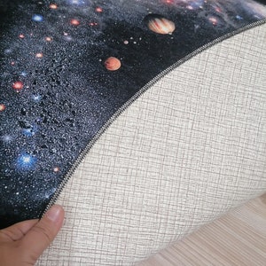 Puede incluir: Una alfombra redonda que presenta una detallada representaci&oacute;n de una galaxia con planetas, estrellas y nebulosas en tonos azules, naranjas y blancos sobre un fondo negro. La parte inferior de la alfombra es de color beige claro con un patr&oacute;n tejido.