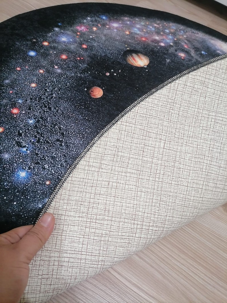 Puede incluir: Una alfombra redonda que presenta una detallada representaci&oacute;n de una galaxia con planetas, estrellas y nebulosas en tonos azules, naranjas y blancos sobre un fondo negro. La parte inferior de la alfombra es de color beige claro con un patr&oacute;n tejido.