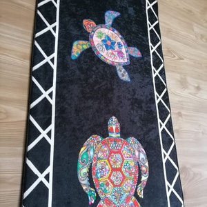 May include: Black rectangular rug featuring two colorful sea turtle designs. The turtles are adorned with intricate patterns in shades of blue, pink, green, and red. A white geometric border frames the rug, adding a decorative touch.