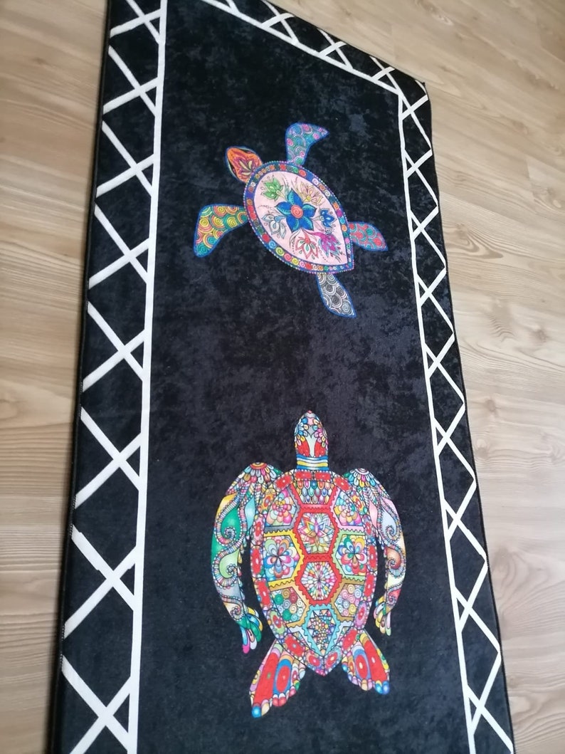 May include: Black rectangular rug featuring two colorful sea turtle designs. The turtles are adorned with intricate patterns in shades of blue, pink, green, and red. A white geometric border frames the rug, adding a decorative touch.