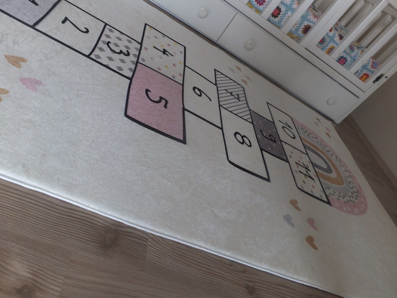 May include: A white play mat with a rainbow design and a hopscotch game printed on it. The hopscotch game features numbers 1 through 10 in black.