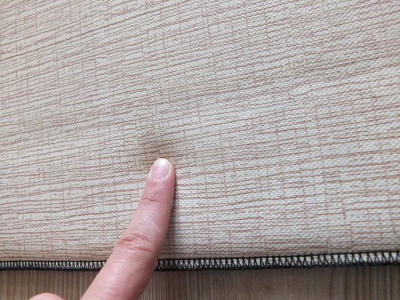 May include: Close-up of a beige fabric with a textured weave. The fabric has a black stitched edge and a small dark spot in the center.