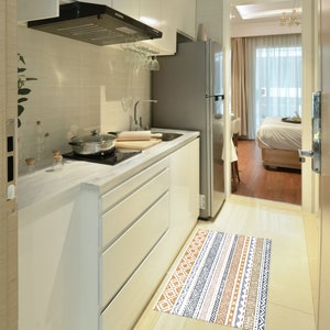May include: A white kitchen with a stainless steel refrigerator, a white countertop, and a black range hood. A patterned rug is on the floor in front of the kitchen.