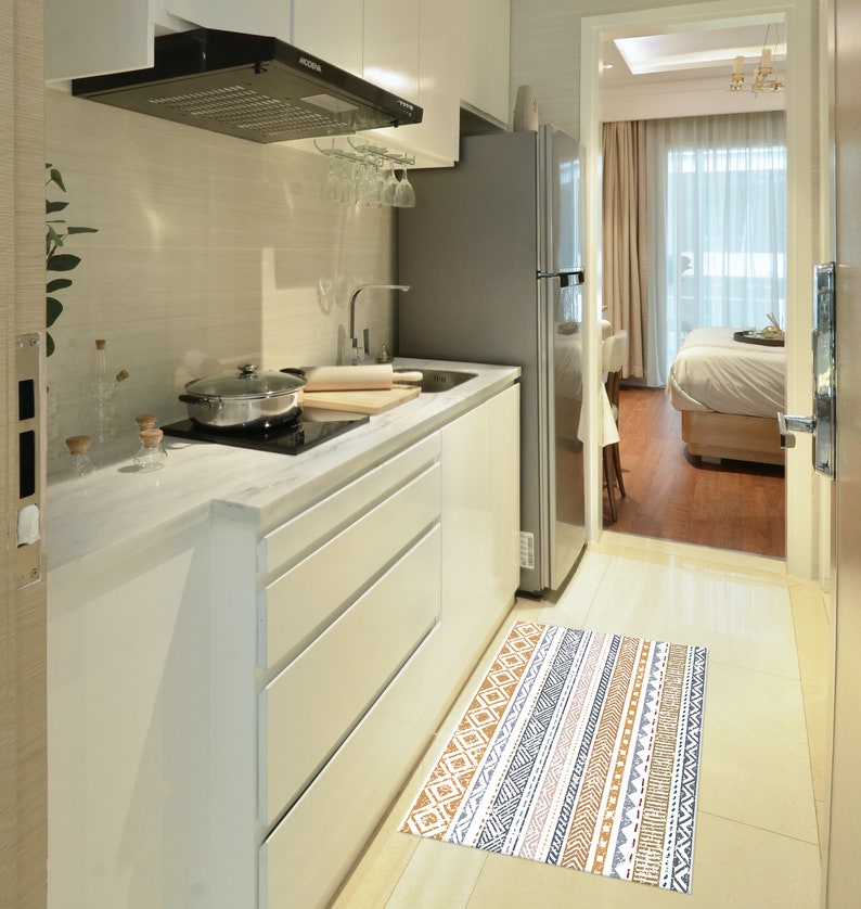May include: A white kitchen with a stainless steel refrigerator, a white countertop, and a black range hood. A patterned rug is on the floor in front of the kitchen.