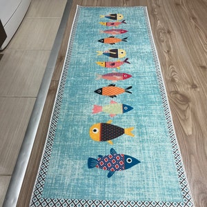 Op de afbeelding: Een blauwe en witte loper met een rand van witte en blauwe geometrische vormen. De loper heeft een rij kleurrijke vissen met zwarte, witte, rode, oranje en gele details.
