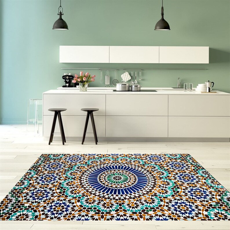K&ouml;nnte beinhalten: Ein farbenfroher Teppich mit geometrischem Muster in einer K&uuml;che. Der Teppich hat einen zentralen blauen Kreis, der von t&uuml;rkisfarbenen, braunen und wei&szlig;en sternf&ouml;rmigen Formen umgeben ist. Die K&uuml;che hat wei&szlig;e Schr&auml;nke, schwarze Hocker und eine hellgr&uuml;ne Wand.