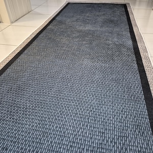 Peut inclure: Un tapis rectangulaire gris foncé avec une texture tissée. Le tapis a une bordure noire et un bord à motifs gris clair et beige. Le tapis est posé sur un sol carrelé blanc.