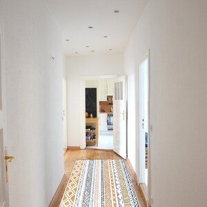 May include: A white hallway with a patterned rug in shades of brown, gray, and white. The rug is laid out in the center of the hallway, leading to an open doorway that reveals a kitchen with white cabinets and a black chalkboard.