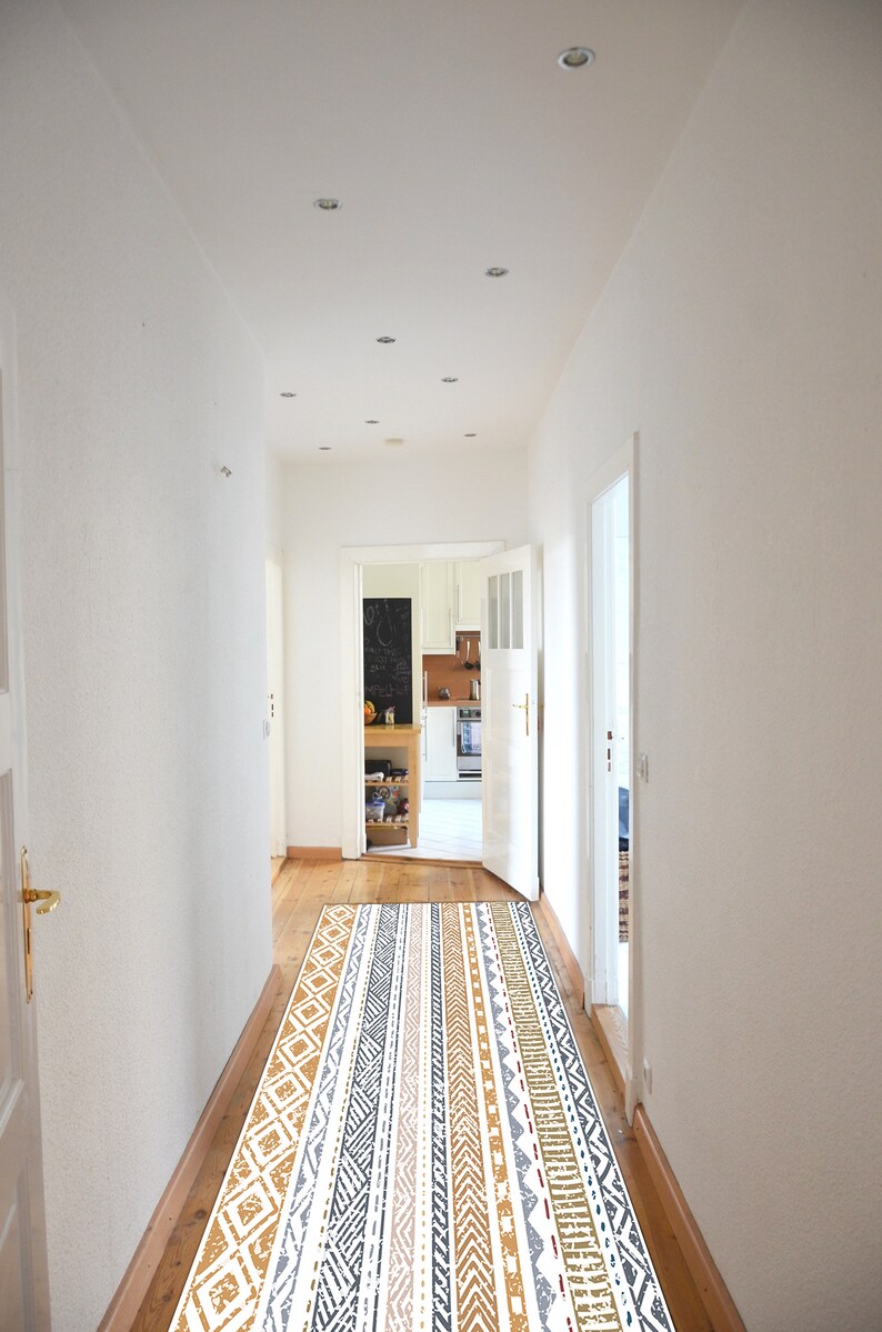 May include: A white hallway with a patterned rug in shades of brown, gray, and white. The rug is laid out in the center of the hallway, leading to an open doorway that reveals a kitchen with white cabinets and a black chalkboard.