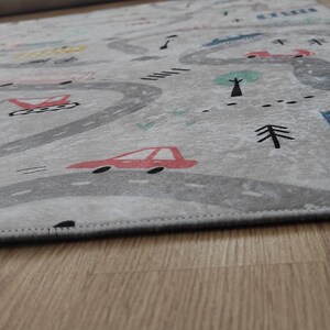 May include: A gray and white play rug with a road design featuring cars, buildings, and trees. The rug has a stitched edge.