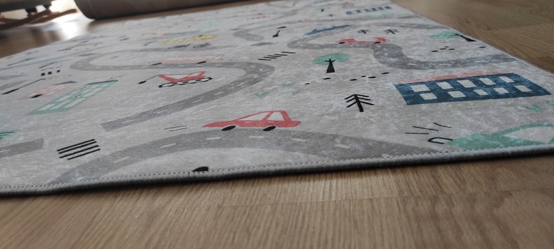 May include: A gray and white play rug with a road design featuring cars, buildings, and trees. The rug has a stitched edge.