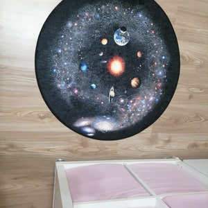 Puede incluir: Una alfombra redonda con una representaci&oacute;n detallada del sistema solar sobre un fondo oscuro. Se ven planetas, estrellas y el sol. La alfombra est&aacute; colocada sobre un suelo de madera clara, con un mueble de almacenamiento blanco y rosa debajo.