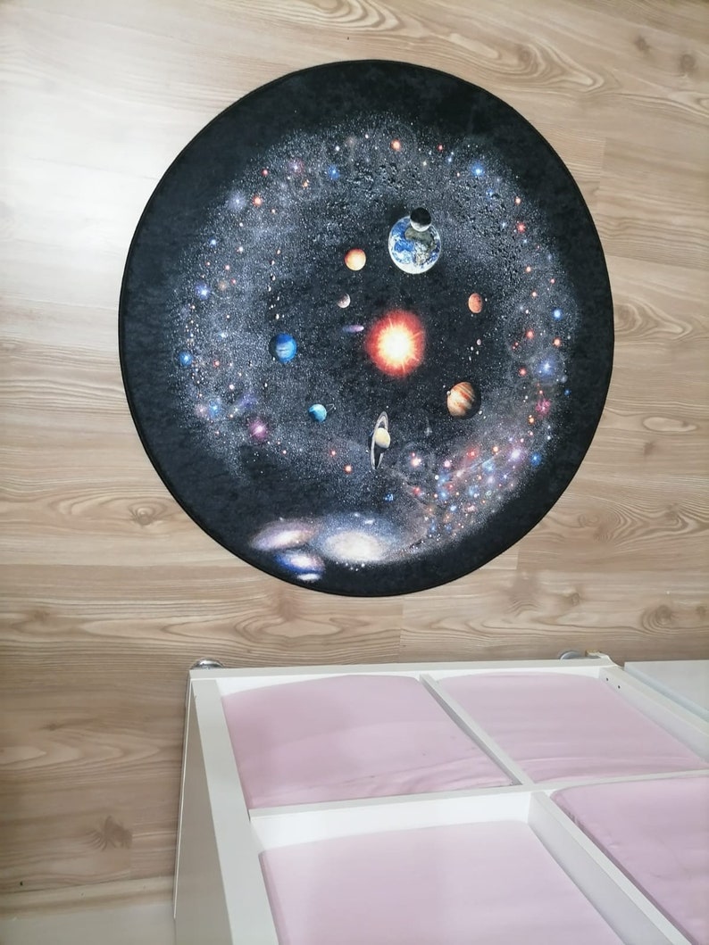 Puede incluir: Una alfombra redonda con una representaci&oacute;n detallada del sistema solar sobre un fondo oscuro. Se ven planetas, estrellas y el sol. La alfombra est&aacute; colocada sobre un suelo de madera clara, con un mueble de almacenamiento blanco y rosa debajo.