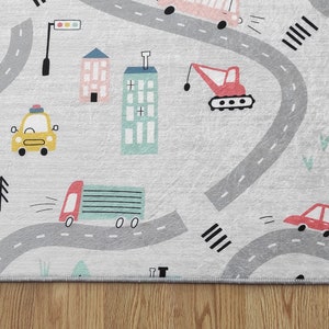 May include: A gray and white play rug with a road and city theme. The rug features a variety of vehicles, including cars, trucks, and a crane. There are also buildings, trees, and traffic lights on the rug.