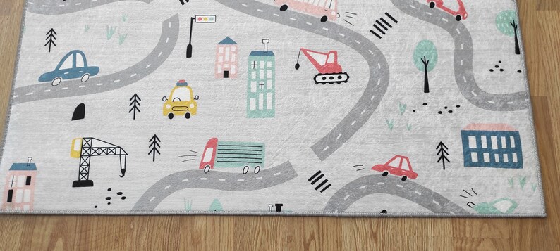 May include: A gray and white play rug with a road and city theme. The rug features a variety of vehicles, including cars, trucks, and a crane. There are also buildings, trees, and traffic lights on the rug.