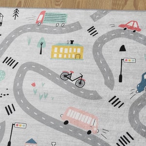 May include: A gray and white play rug with a city theme. The rug features a winding road with cars, a bus, a bicycle, a crane, and buildings. The rug is perfect for imaginative play.