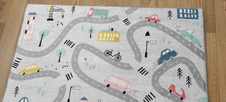 May include: A gray and white play rug with a city theme. The rug features a winding road with cars, a bus, a bicycle, a crane, and buildings. The rug is perfect for imaginative play.