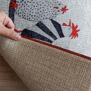 May include: A grey fabric featuring a repeating pattern of stylised chickens in black, white, and red. The underside of the fabric is tan with a red border. A hand is holding the fabric.
