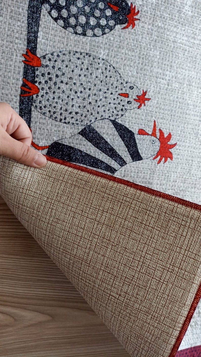 May include: A grey fabric featuring a repeating pattern of stylised chickens in black, white, and red. The underside of the fabric is tan with a red border. A hand is holding the fabric.