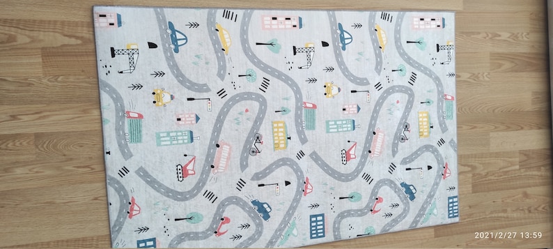 May include: A gray and white play mat with a city theme. The mat features a winding road with cars, trucks, and buildings. The road is surrounded by trees and streetlights.
