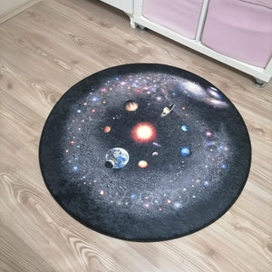 Puede incluir: Una alfombra redonda que muestra una detallada representaci&oacute;n del sistema solar sobre un fondo oscuro y estrellado. Se ven planetas, el sol y galaxias. La alfombra est&aacute; colocada sobre un suelo de madera clara, lo que sugiere un tema decorativo o educativo.