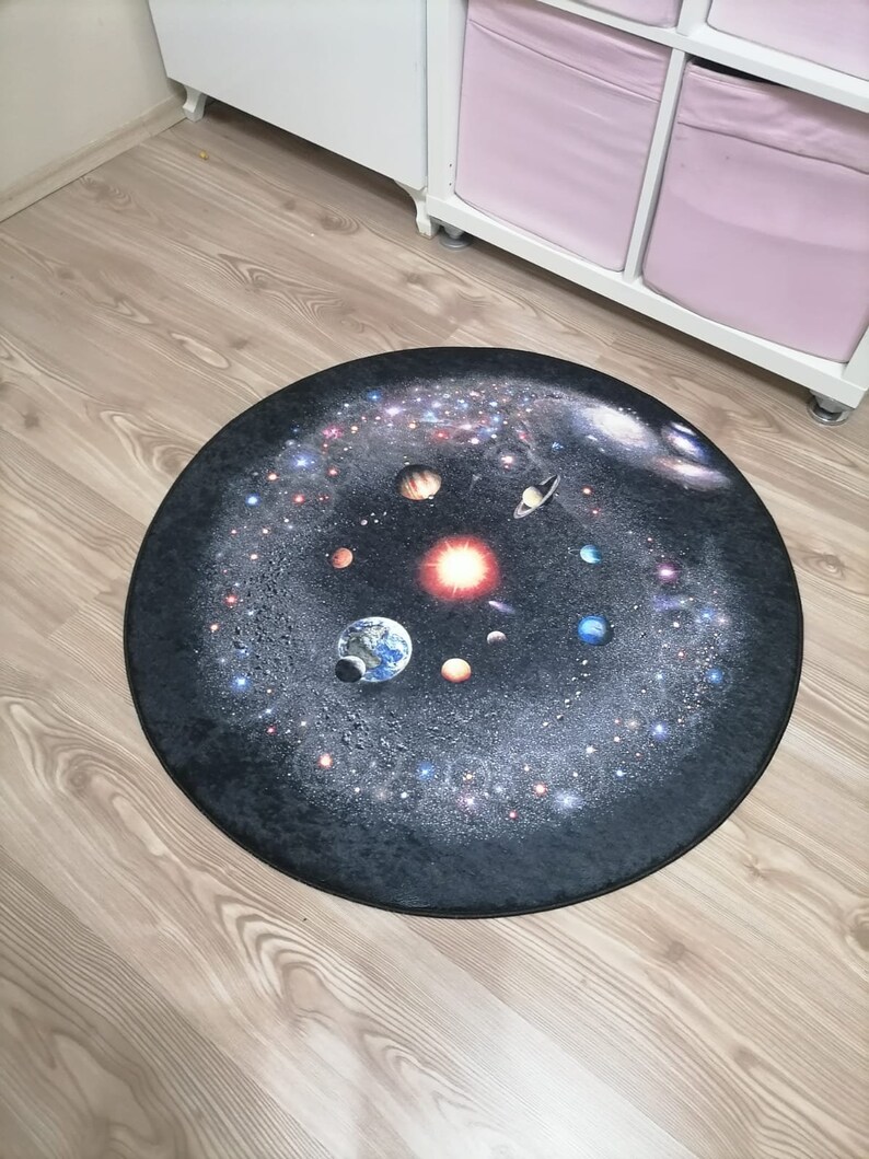 Puede incluir: Una alfombra redonda que muestra una detallada representaci&oacute;n del sistema solar sobre un fondo oscuro y estrellado. Se ven planetas, el sol y galaxias. La alfombra est&aacute; colocada sobre un suelo de madera clara, lo que sugiere un tema decorativo o educativo.
