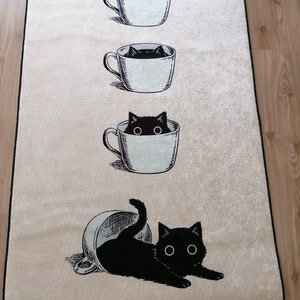 May include: A white rug with a black cat peeking out of a coffee cup. The cat is in three different stages of being in the cup, starting with just its head, then its head and shoulders, and finally the whole cat. The rug has a beige background.