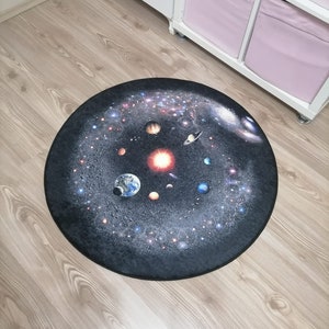 Puede incluir: Una alfombra redonda que presenta una representaci&oacute;n detallada del sistema solar sobre un fondo oscuro. Los planetas, las estrellas y las galaxias son visibles en esta alfombra con tem&aacute;tica espacial, colocada sobre un suelo de madera.