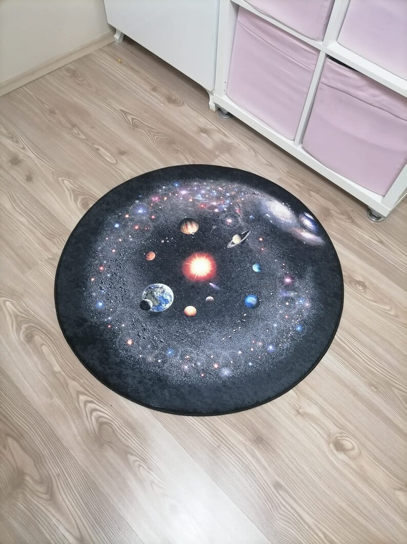 Puede incluir: Una alfombra redonda que presenta una representaci&oacute;n detallada del sistema solar sobre un fondo oscuro. Los planetas, las estrellas y las galaxias son visibles en esta alfombra con tem&aacute;tica espacial, colocada sobre un suelo de madera.