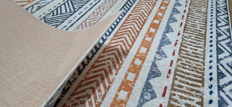 May include: A close-up of a beige and white patterned rug with geometric designs in shades of blue, orange, and brown. The rug is folded over, revealing the beige backing.