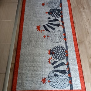 May include: Rectangular grey rug with a red border. The rug features a repeating pattern of stylised chickens in black and white, perched on a vertical line. The chickens have various patterns, including polka dots and stripes. The rug is on a wooden floor.