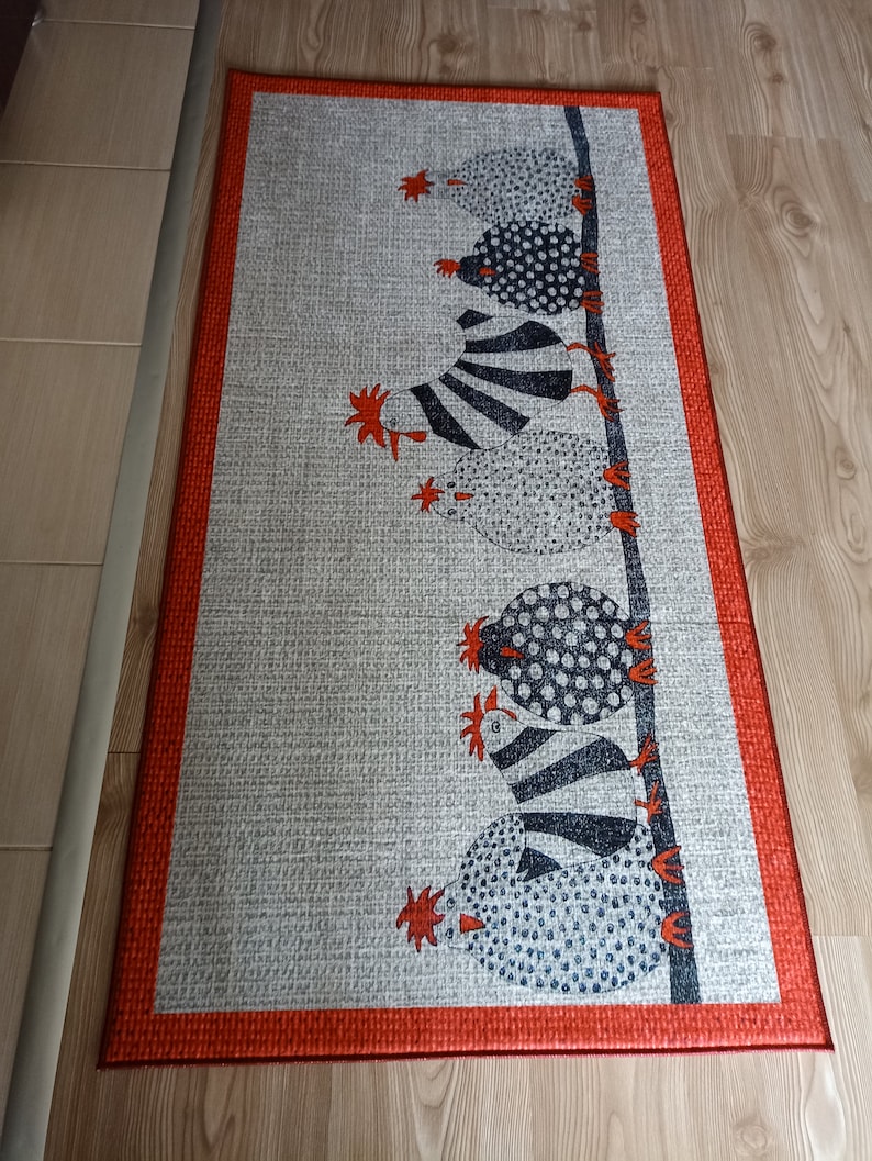 May include: Rectangular grey rug with a red border. The rug features a repeating pattern of stylised chickens in black and white, perched on a vertical line. The chickens have various patterns, including polka dots and stripes. The rug is on a wooden floor.