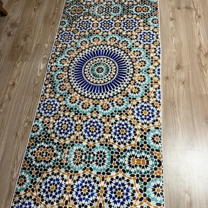 K&ouml;nnte beinhalten: Ein rechteckiger Teppich mit einem detaillierten geometrischen Muster in Blau-, T&uuml;rkis-, Gold- und Schwarzt&ouml;nen. Das Design weist ein zentrales kreisf&ouml;rmiges Motiv auf, das von komplizierten sternartigen Formen umgeben ist. Der Teppich hat einen wei&szlig;en Rand und liegt auf einem Holzboden.