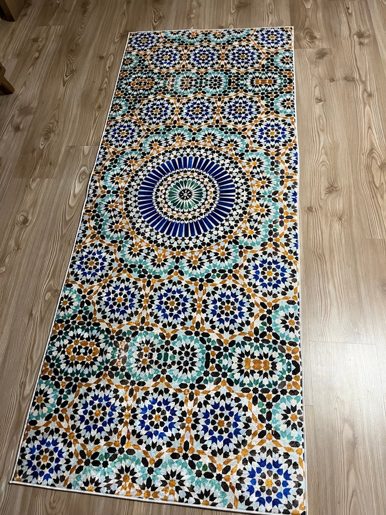 K&ouml;nnte beinhalten: Ein rechteckiger Teppich mit einem detaillierten geometrischen Muster in Blau-, T&uuml;rkis-, Gold- und Schwarzt&ouml;nen. Das Design weist ein zentrales kreisf&ouml;rmiges Motiv auf, das von komplizierten sternartigen Formen umgeben ist. Der Teppich hat einen wei&szlig;en Rand und liegt auf einem Holzboden.