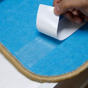 K&ouml;nnte beinhalten: Ein blauer Teppich mit einem beigefarbenen Fransenrand ist mit einem durchsichtigen Klebeband auf der R&uuml;ckseite versehen. Eine Hand zieht die wei&szlig;e Papierunterlage vom Klebeband ab.