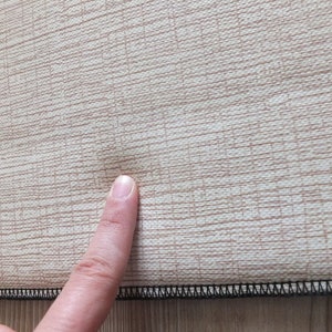 Puede incluir: Primer plano de una tela beige con un patr&oacute;n de tejido texturizado. La tela tiene una sutil variaci&oacute;n de color natural. Un dedo toca la tela, resaltando una peque&ntilde;a imperfecci&oacute;n. El borde es negro.