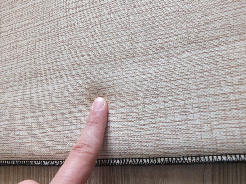 Puede incluir: Primer plano de una tela beige con un patr&oacute;n de tejido texturizado. La tela tiene una sutil variaci&oacute;n de color natural. Un dedo toca la tela, resaltando una peque&ntilde;a imperfecci&oacute;n. El borde es negro.