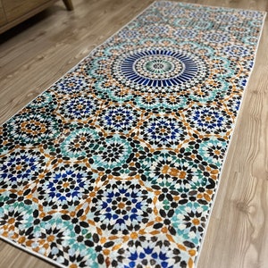 K&ouml;nnte beinhalten: Rechteckiger Teppich mit detailliertem geometrischen Muster in Blau-, T&uuml;rkis-, Braun- und Wei&szlig;t&ouml;nen. Das Design weist ein zentrales kreisf&ouml;rmiges Motiv und sich wiederholende sternartige Formen auf, die einen Mosaikeffekt erzeugen. Der Teppich liegt auf einem Holzboden.