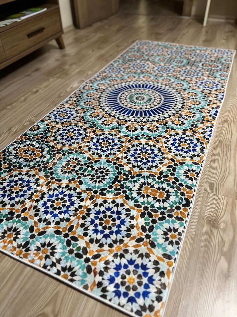 K&ouml;nnte beinhalten: Rechteckiger Teppich mit detailliertem geometrischen Muster in Blau-, T&uuml;rkis-, Braun- und Wei&szlig;t&ouml;nen. Das Design weist ein zentrales kreisf&ouml;rmiges Motiv und sich wiederholende sternartige Formen auf, die einen Mosaikeffekt erzeugen. Der Teppich liegt auf einem Holzboden.