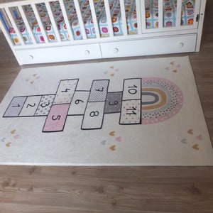 May include: A white rug with a hopscotch design featuring numbers 1 through 11. The rug also has a rainbow with pink, yellow, and gray stripes and a scattering of pink and gold hearts.