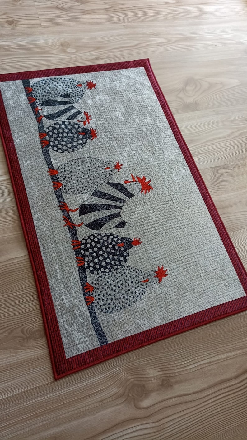 May include: Rectangular rug with a light grey background and a dark red border. The rug features a design of stylized chickens in black and white with red accents, perched on a dark grey branch. Suitable for a kitchen or entryway.