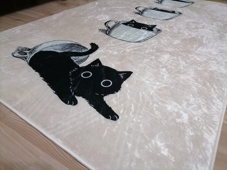 May include: A white rug with a black cat design. The cat is peeking out of a teacup. The rug has a soft, velvety texture.